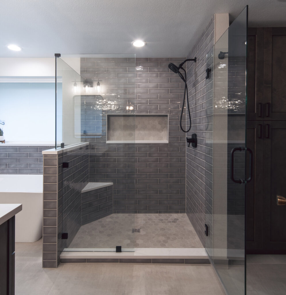 A bathroom remodel displaying a large shower featuring glossy grey subway tile for the surround, a corner bench with a quartz top, and hexagonal tile for the shower pan. There are black shower fixtures. 