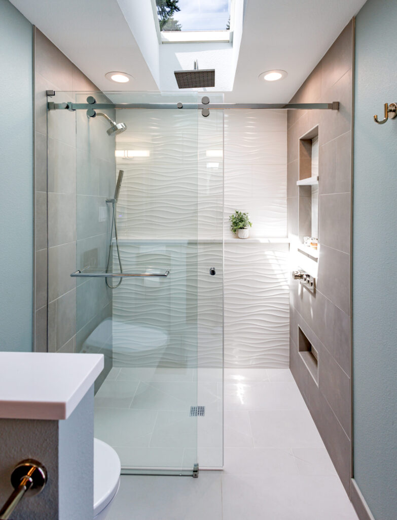 A Mill Creek Bathroom Remodel featuring A large shower with wave pattern tile along the back, large grey tile on either side, polished stainless fixtures, and a quartz shelf along the back wall. There is a skylight above the top of the shower letting in natural light, along with a rain head in the middle of the skylight. 