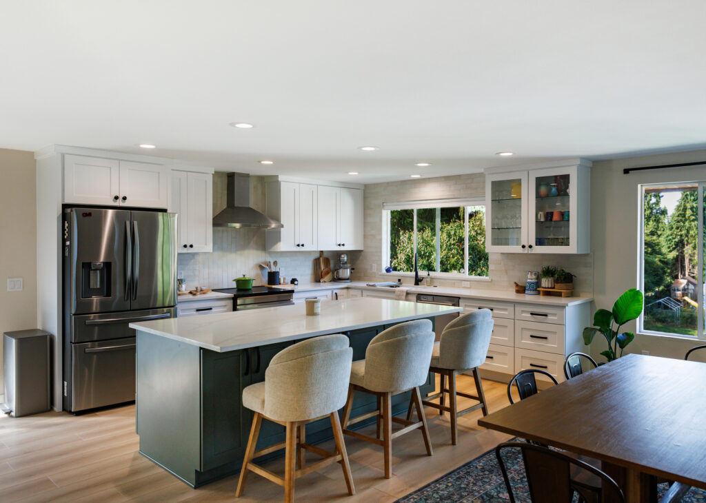 Open-concept kitchen remodel with a green center island, a glass-front upper cabinet, quartz countertops, and a large window overlooking a wooded backyard.