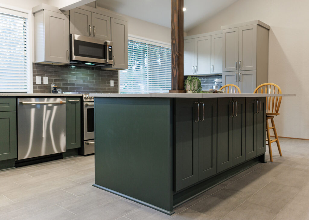 Green kitchen island with quartz countertop, brushed nickel hardware, and a wood support post, paired with gray perimeter cabinetry and gray tile floors.